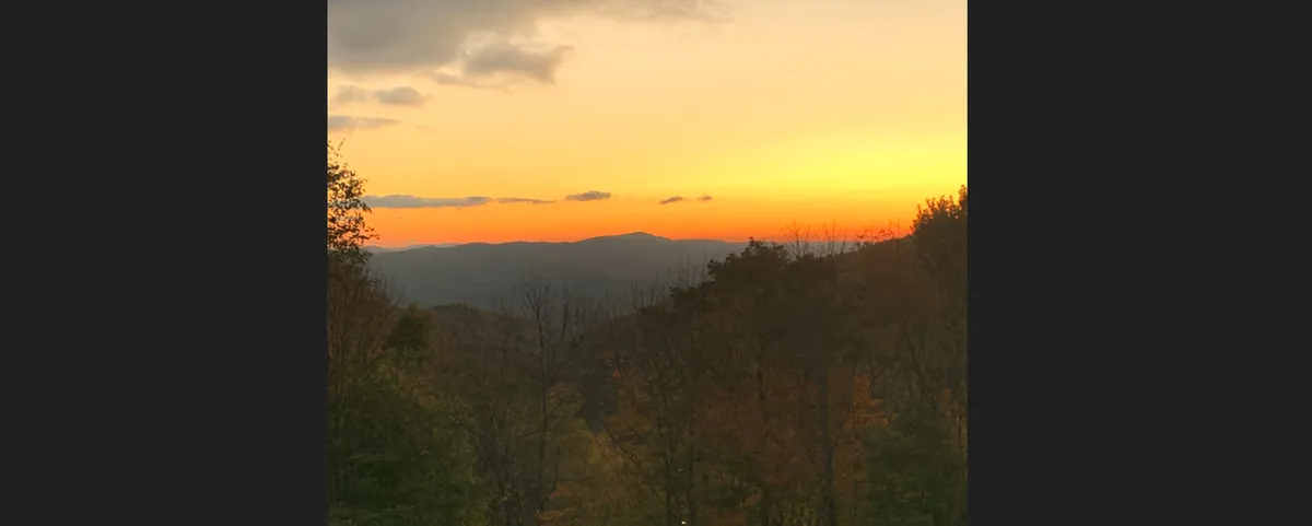 The image showcases a sweeping view of the Blue Ridge Mountains at sunset from the property's vantage point. Warm, golden light bathes the distant peaks, with lush foliage framing the scene. The tranquil atmosphere invites relaxation and quiet appreciation of nature's beauty.