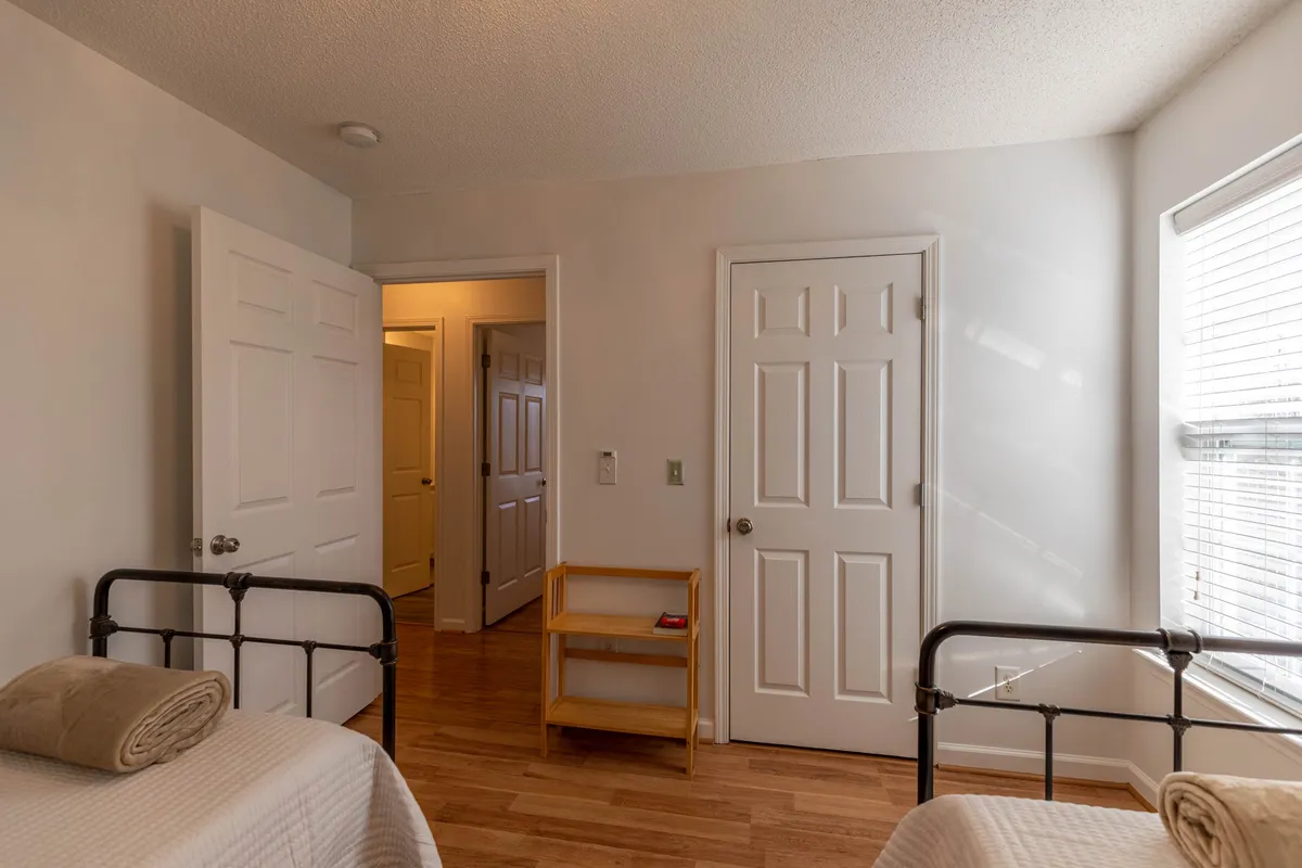 A serene bedroom scene with metal bed frames, a wooden shelf, and windows with blinds. The scene depicts a tranquil spot for guests to relax, read, or savor the morning light streaming in, further suggesting a connection to the surrounding natural beauty.