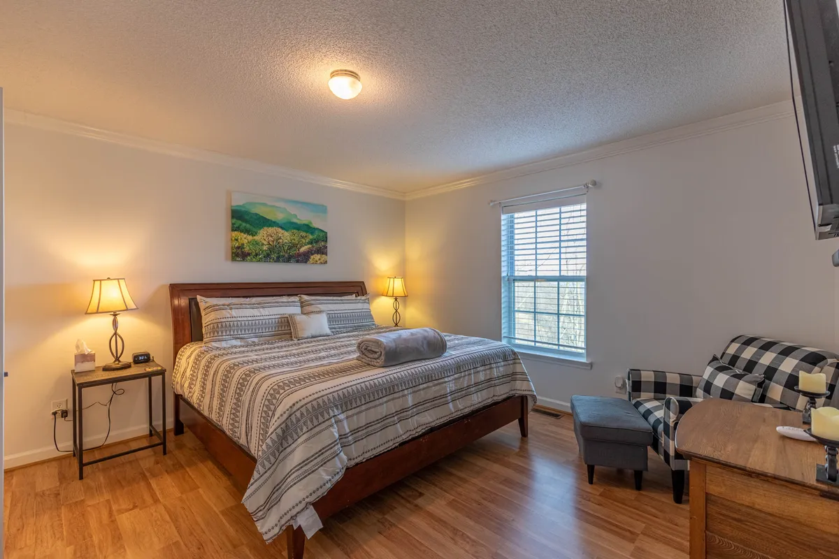 This image features a cozy bedroom with a comfortable bed, a lamp, window, artwork, and a chair. The window allows for views of the outdoors and gives the impression of a space where guests could watch the sunrise or sip coffee while admiring the mountains.