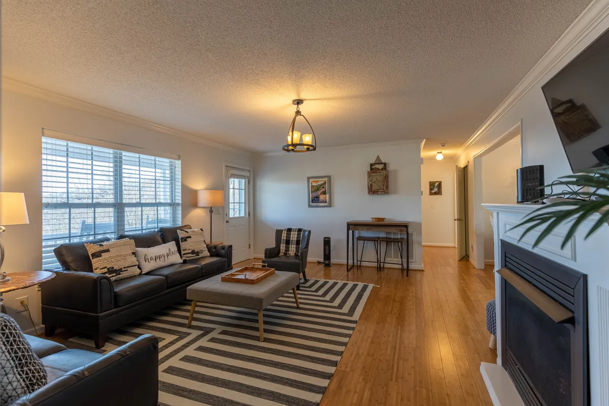 A welcoming great room featuring a plush sofa, a coffee table, and a centerpiece gas log fireplace. Large windows let in an abundance of natural light, enhancing the inviting atmosphere. Artwork on the walls and a glimpse of the outdoors through the window help create a cozy yet scenic living space.