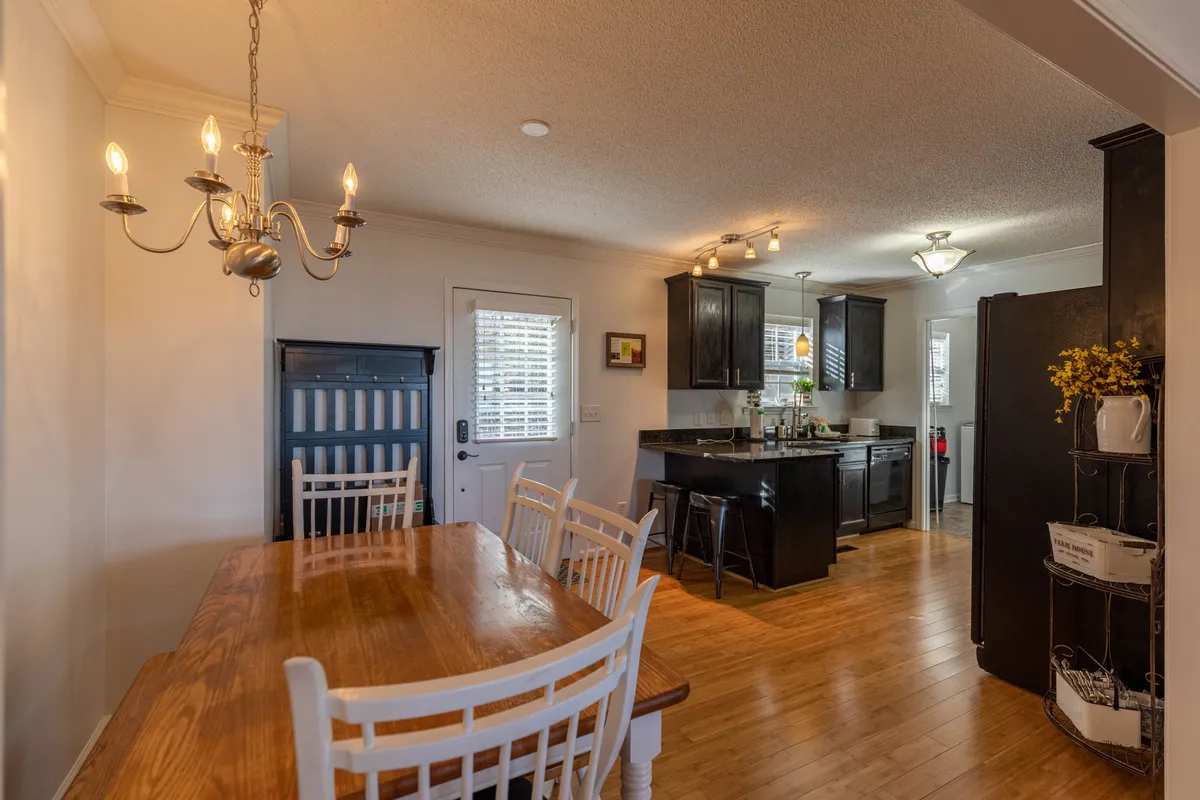 This image features a dining area with a sturdy dining table, a warm chandelier overhead, and a visible kitchen counter, with a doorway leading to other parts of the cabin. The space feels communal and connected, echoing the gathering and sharing aspect of a mountain retreat.