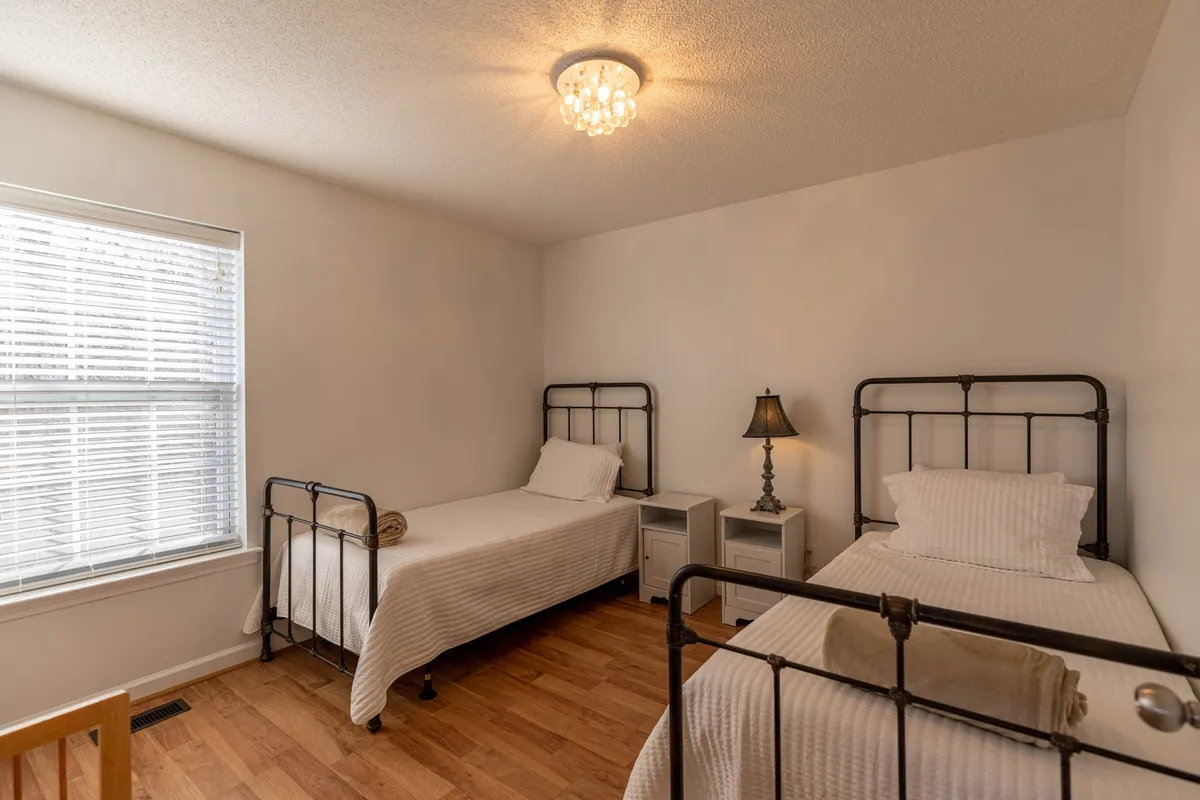 A cozy bedroom with metal bed frames, a nightstand with a lamp, and a generous window with blinds. The ambiance is tranquil and inviting, suitable for guests or family members needing a restful retreat.
